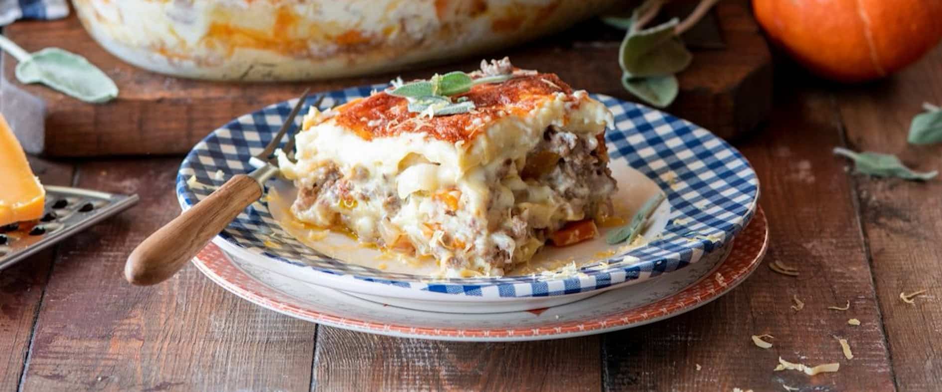 Kürbis-Lasagne mit Hackfleisch, cremiger Sauce und geschmolzenem Käse, serviert auf einem Teller, im Hintergrund eine Auflaufform mit frischer Lasagne und Herbstdeko mit Kürbissen.
