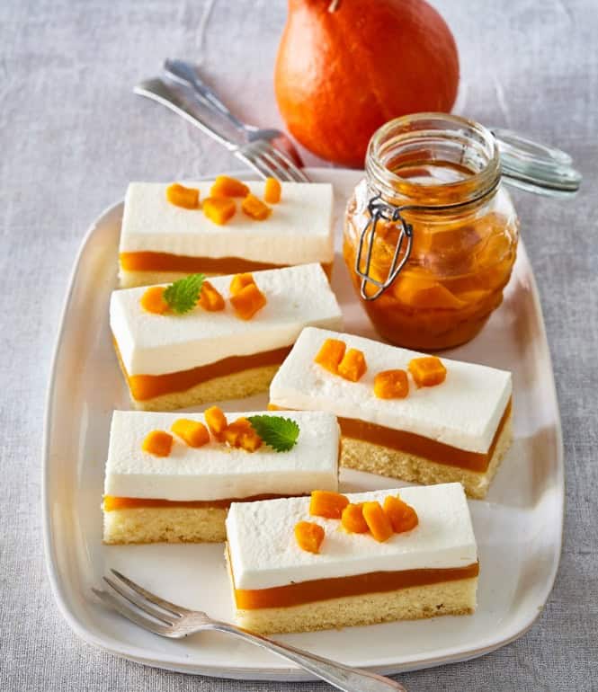Kürbis-Schnitten mit luftigem Sahne-Quark-Topping, Kürbispudding-Schicht und Biskuitboden, garniert mit Kürbiswürfeln, auf einer Servierplatte angerichtet.