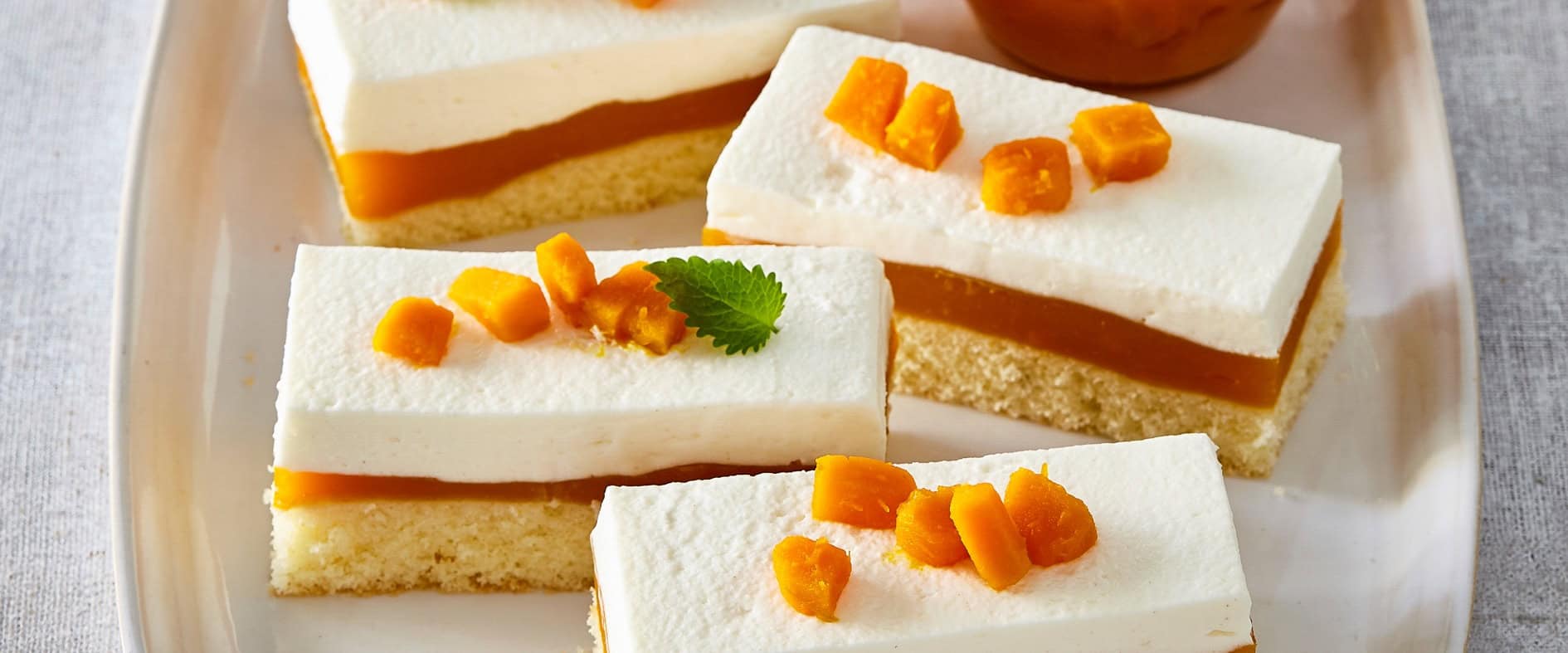 Kürbis-Schnitten mit luftigem Sahne-Quark-Topping, Kürbispudding-Schicht und Biskuitboden, garniert mit Kürbiswürfeln, auf einer Servierplatte angerichtet.