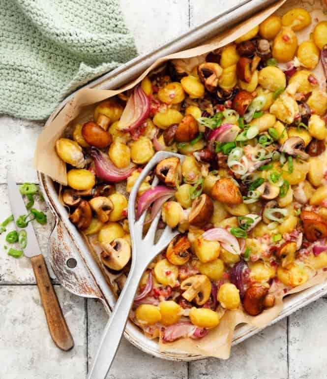 Ofen-Gnocchi mit gebratenen Champignons, roten Zwiebeln, Speckwürfeln und geschmolzenem Käse, garniert mit Frühlingszwiebeln in einer Auflaufform.