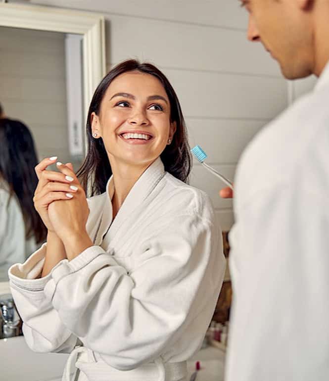 Paar in weißen Bademänteln lächelt sich im Badezimmer an.