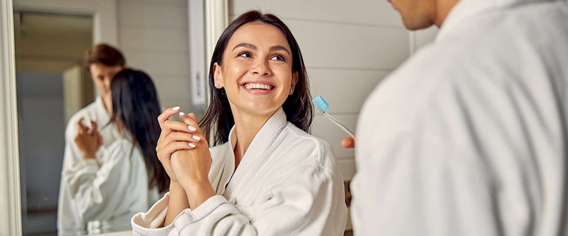 Paar in weißen Bademänteln lächelt sich im Badezimmer an.