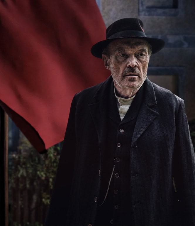 Josef Hader als Julius Kraus in der ZDF-Historienserie „Sturm kommt auf“, stehend vor einem Gebäude mit roter Fahne.