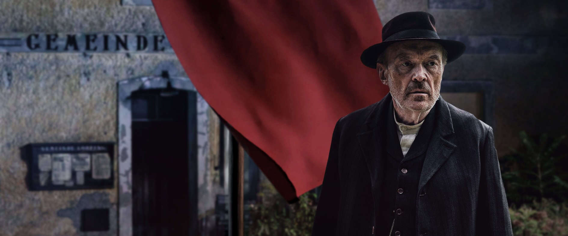 Josef Hader als Julius Kraus in der ZDF-Historienserie „Sturm kommt auf“, stehend vor einem Gebäude mit roter Fahne.