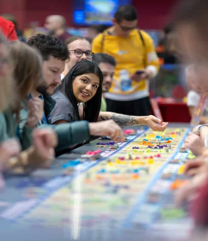 Mehrere Menschen sitzen an einem langen Tisch und spielen Catan mit vielen bunten Spielfiguren auf der Messe SPIEL in Essen.