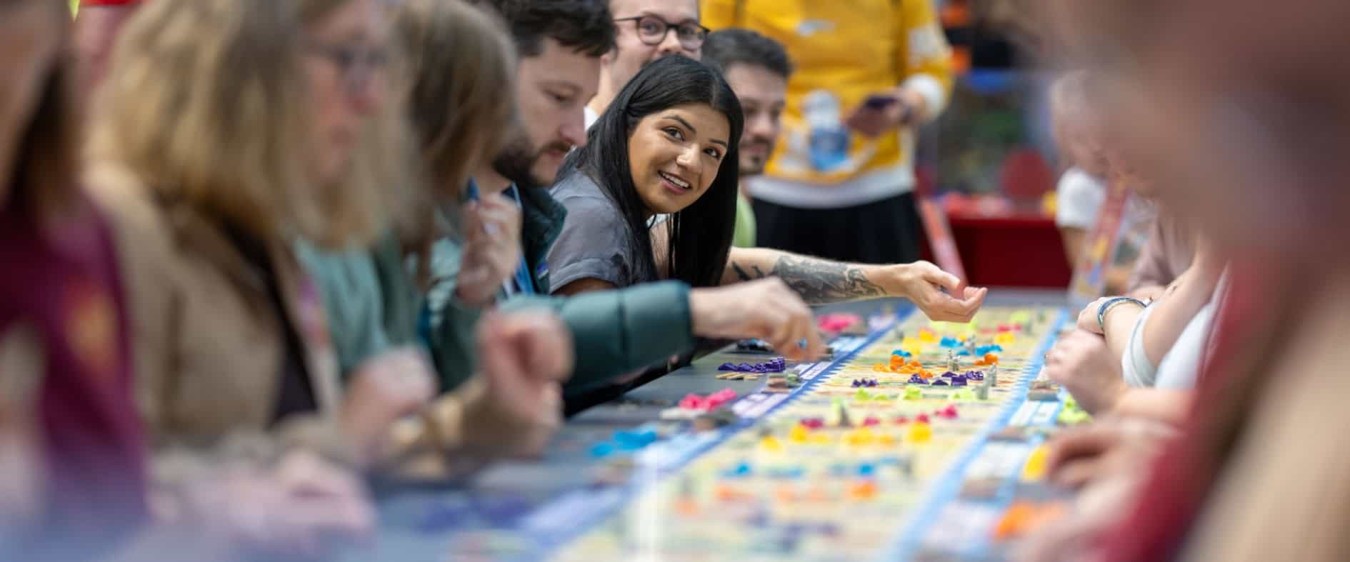 Mehrere Menschen sitzen an einem langen Tisch und spielen Catan mit vielen bunten Spielfiguren auf der Messe SPIEL in Essen.
