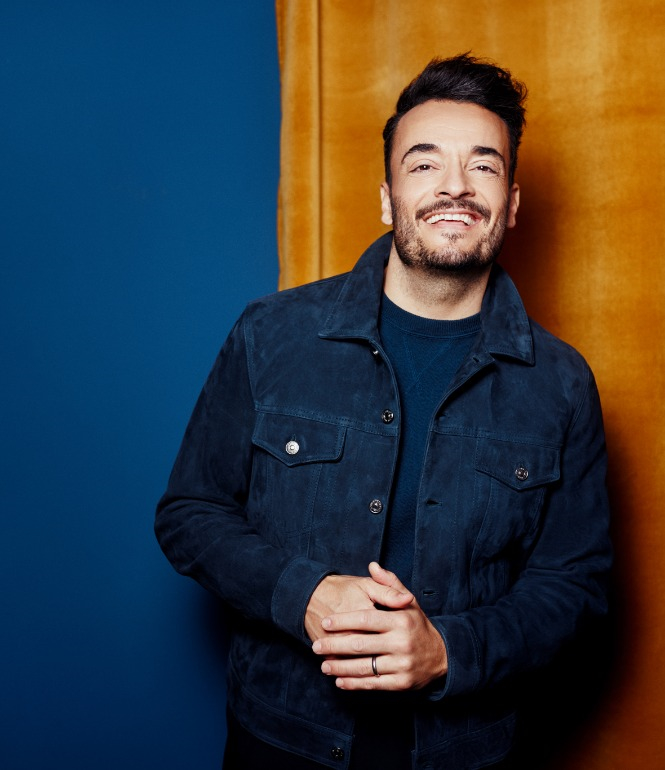 Giovanni Zarella lächelt in dunkelblauer Jacke vor blauem und gelbem Studiohintergrund, in entspannter Pose.