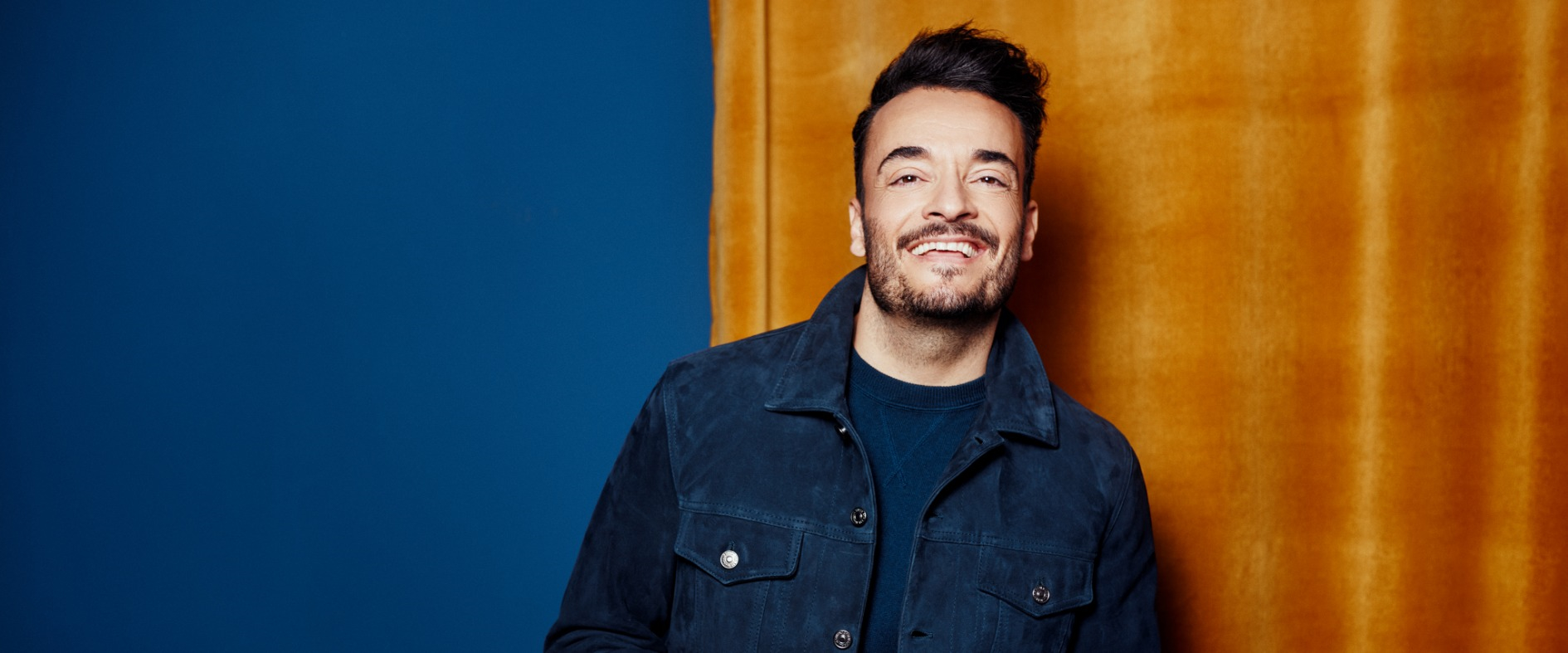 Giovanni Zarella lächelt in dunkelblauer Jacke vor blauem und gelbem Studiohintergrund, in entspannter Pose.