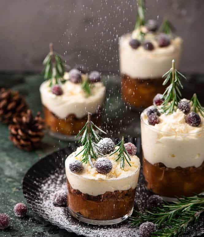 Winterliches Bratapfel-Tiramisu im Glas, geschichtet mit Mascarponecreme, Lebkuchen und dekoriert mit Rosmarinzweigen und gezuckerten Beeren, leicht mit Puderzucker bestäubt.