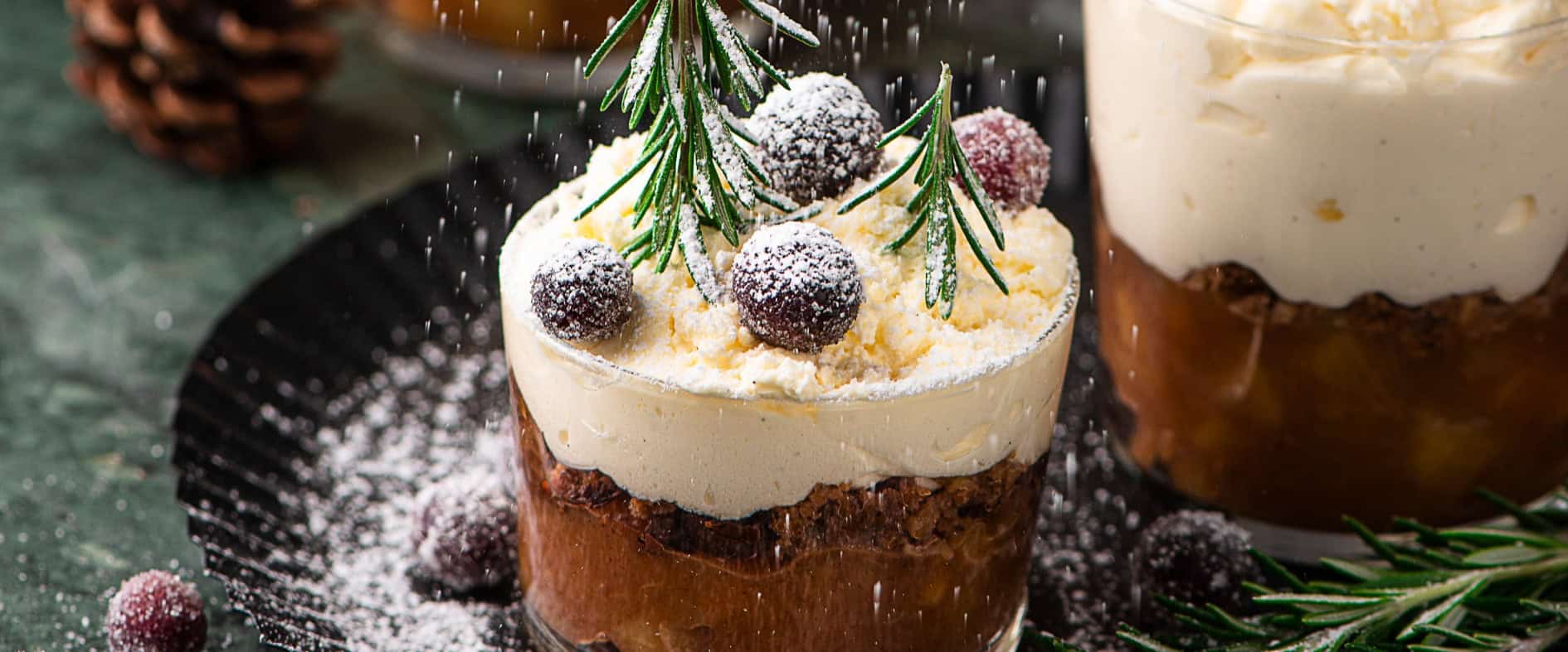 Winterliches Bratapfel-Tiramisu im Glas, geschichtet mit Mascarponecreme, Lebkuchen und dekoriert mit Rosmarinzweigen und gezuckerten Beeren, leicht mit Puderzucker bestäubt.