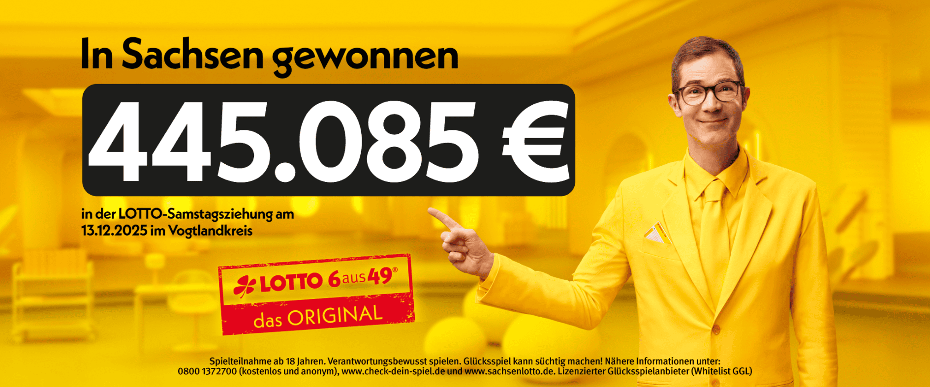 Ein freundlich lächelnder Mann in gelbem Anzug zeigt auf einen Gewinn von 445.085 Euro bei der LOTTO-Samstagsziehung am 13.12.2025 im Vogtlandkreis.