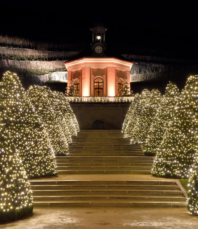 Weihnachtlich beleuchtete Gartenanlage von Schloss Wackerbarth mit Lichterbäumen und barocker Turmfassade im Hintergrund
