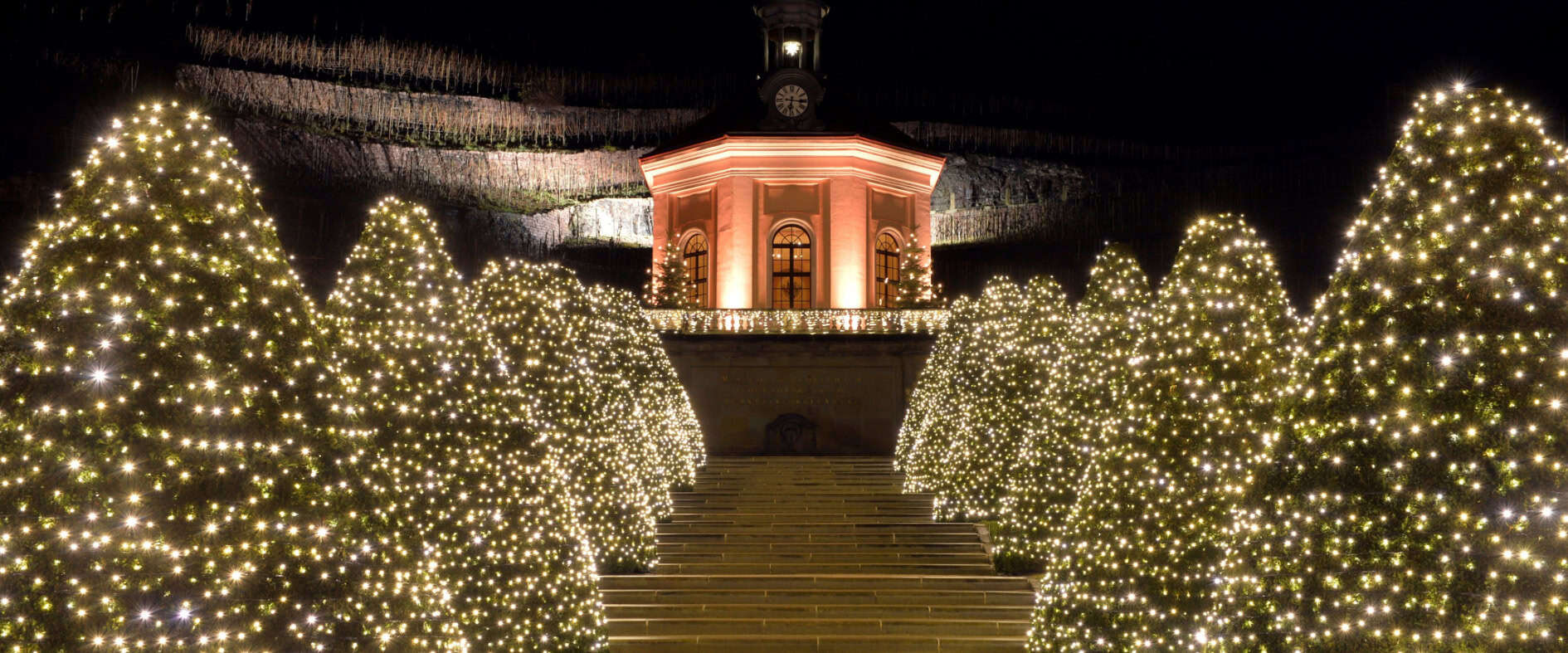 Weihnachtlich beleuchtete Gartenanlage von Schloss Wackerbarth mit Lichterbäumen und barocker Turmfassade im Hintergrund