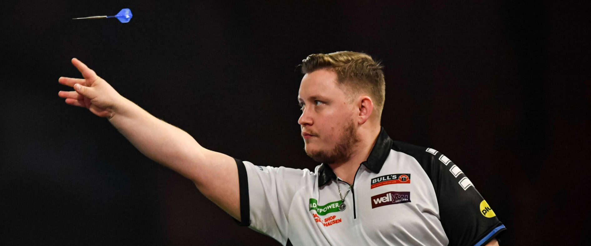 Dartspieler Martin Schindler wirft einen Pfeil präzise in Richtung Scheibe, fokussiert und in Wettkampfatmosphäre.