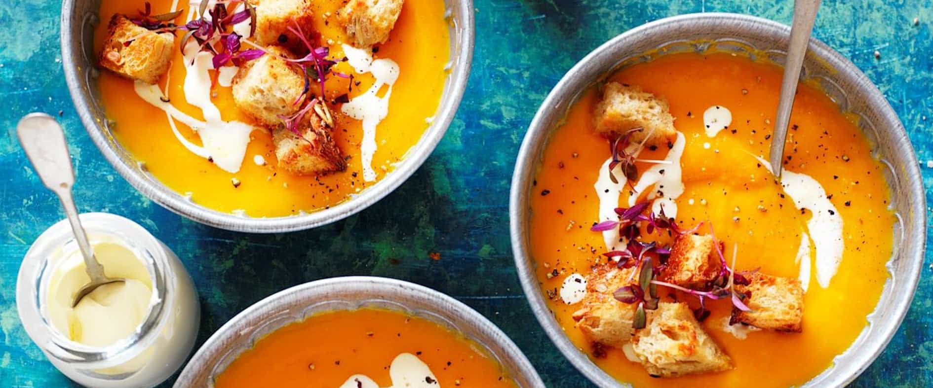 Cremige Süßkartoffel-Kürbis-Suppe in drei Schalen, serviert mit knusprigen Toast-Croutons, einem Klecks Saurer Sahne und frischen Kräutern.