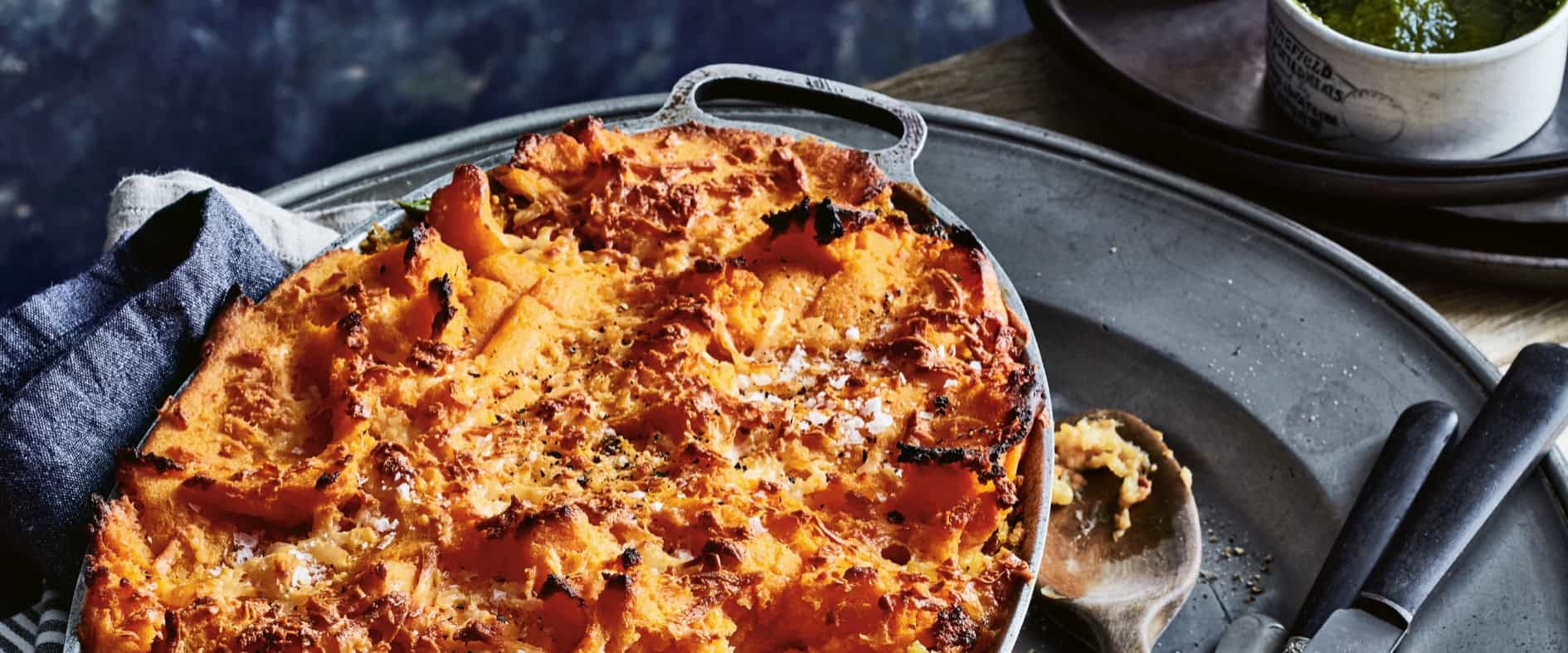 Vegetarischer Shepherd’s Pie aus Kartoffeln, Süßkartoffeln und Gemüse-Reis, goldbraun überbacken in einer Ofenform, serviert rustikal mit Besteck und Pesto.