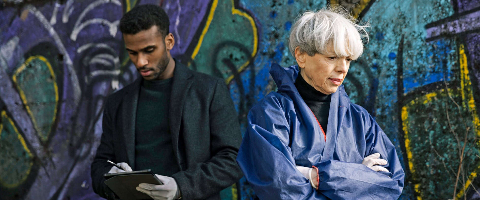 Zwei Ermittler:innen stehen nachdenklich vor einer Graffitiwand.