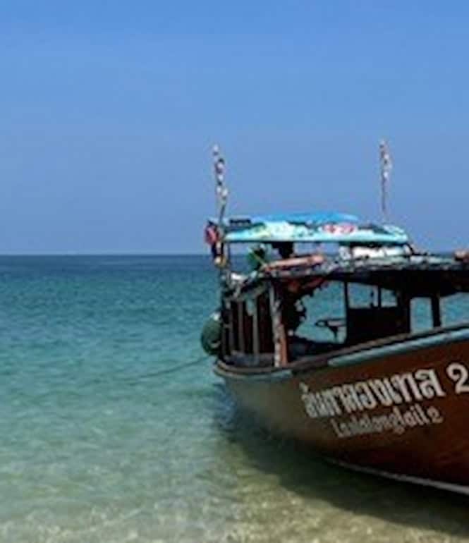 Ein traditionelles thailändisches Langboot (Longtail-Boot) mit bunter Bemalung und Schmuck liegt am flachen, sandigen Strand eines türkisblauen Meeres unter klarem Himmel.