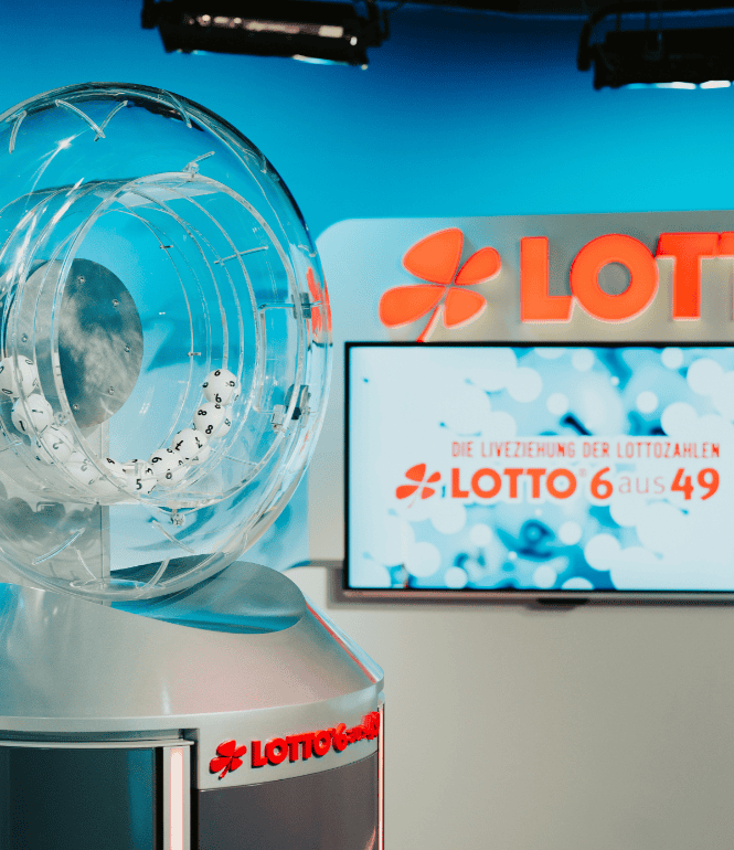 Transparente Lotto-Ziehungstrommel mit weißen Zahlenkugeln während einer LOTTO-6aus49-Ziehung, im Hintergrund das LOTTO-Logo und ein Bildschirm mit der Aufschrift „Die Liveziehung der Lottozahlen“.