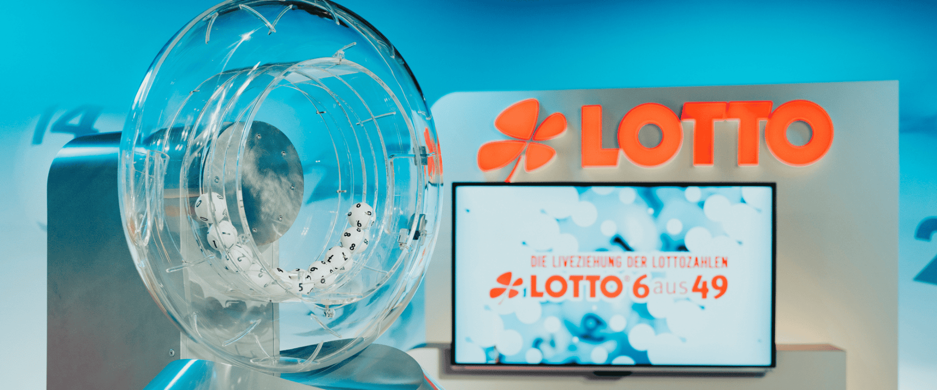Transparente Lotto-Ziehungstrommel mit weißen Zahlenkugeln während einer LOTTO-6aus49-Ziehung, im Hintergrund das LOTTO-Logo und ein Bildschirm mit der Aufschrift „Die Liveziehung der Lottozahlen“.