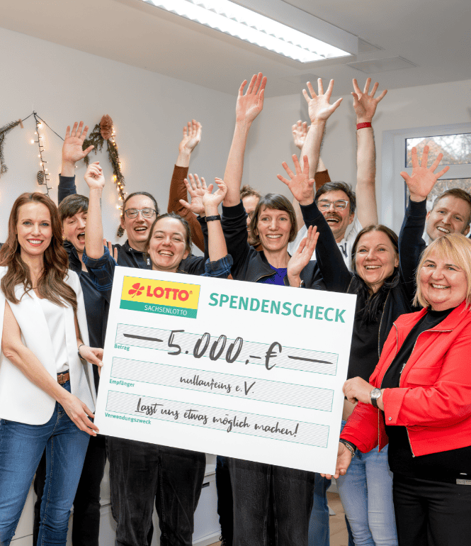 Gruppenfoto bei einer symbolischen Scheckübergabe: Susanne Schöne überreicht im Namen von Sachsenlotto 5.000 Euro an den Leipziger Verein nullaufeins e.V. – umgeben von fröhlichen Vereinsmitgliedern, die gemeinsam feiern.