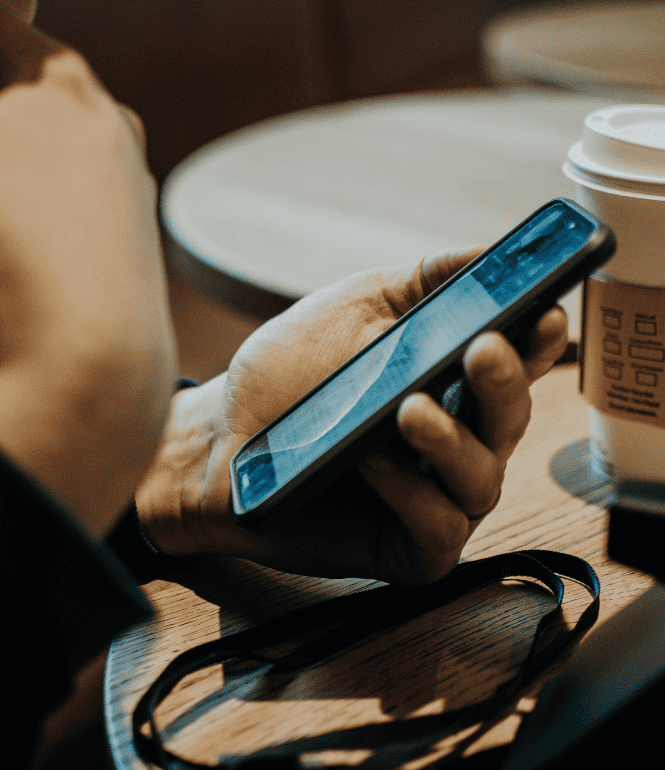Person hält Smartphone in der Hand, im Hintergrund ein Kaffeebecher – Symbolbild für Betrugsanrufe