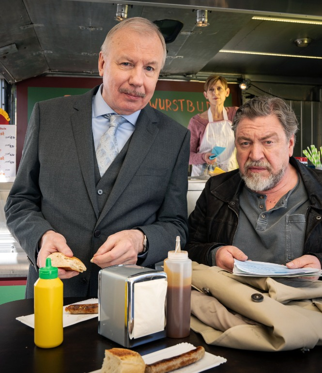 Zwei Männer essen Bratwurst an einem Stehtisch vor einer Imbissbude, im Hintergrund eine Verkäuferin im Verkaufswagen