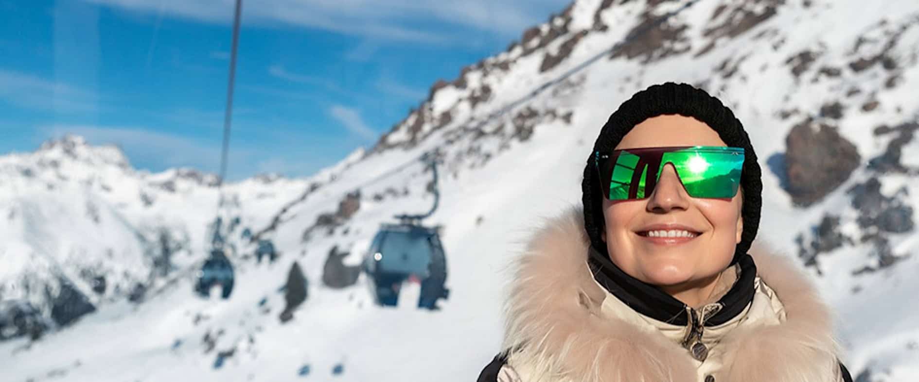 Frau in luxuriöser Winterkleidung mit verspiegelter Skibrille vor verschneitem Alpenpanorama und Gondelbahn