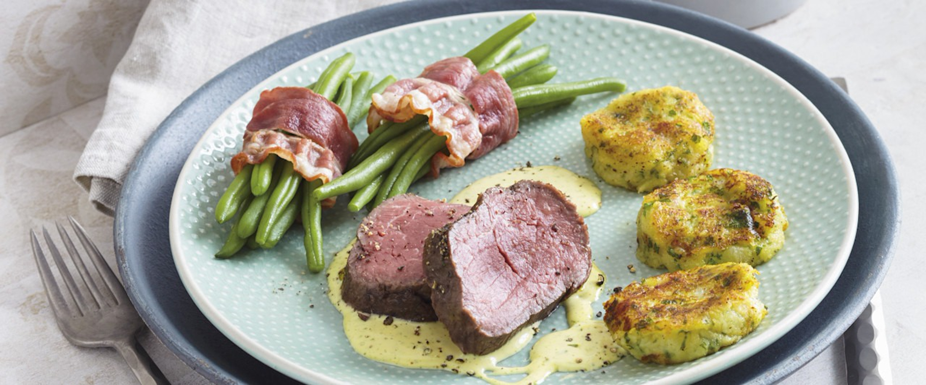 Zartrosa gebratenes Rinderfilet mit Speckbohnen, goldbraunen Macaire-Kartoffeln und cremiger Estragon-Sauce auf einem Teller angerichtet.