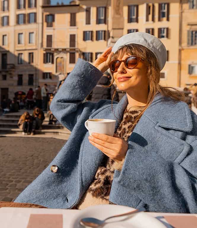 Frau sitzt mit einer Tasse Kaffee in der Sonne auf einem belebten Platz und genießt den Moment.