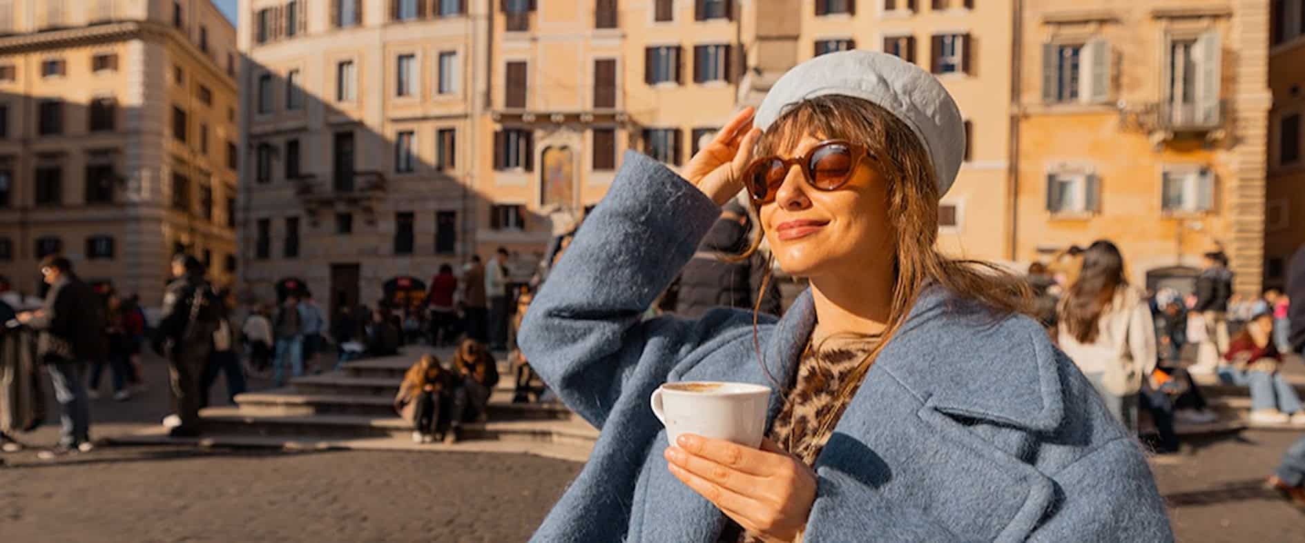 Frau sitzt mit einer Tasse Kaffee in der Sonne auf einem belebten Platz und genießt den Moment.