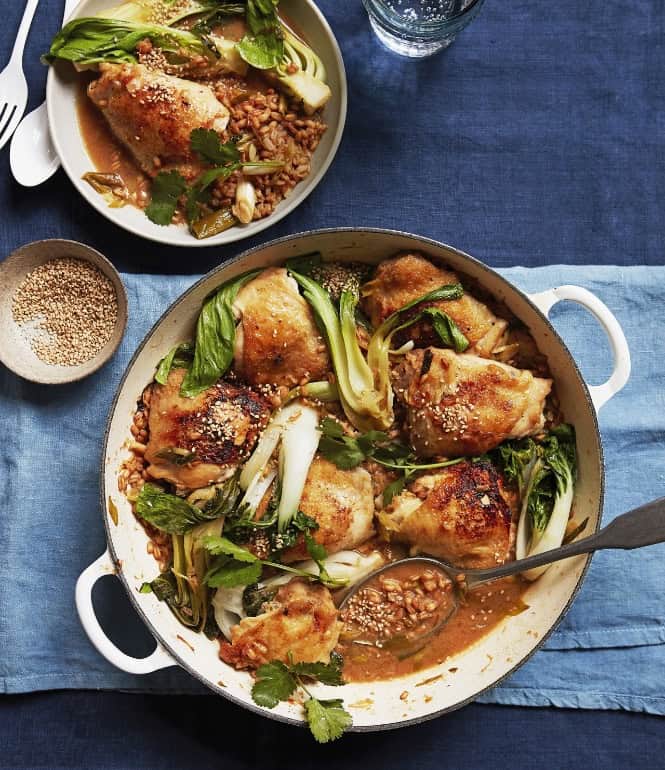 One-Pot-Huhn mit Pak Choi und Perldinkel in einer Pfanne, garniert mit Sesam und frischem Koriander, serviert mit einer Schüssel und Löffel.