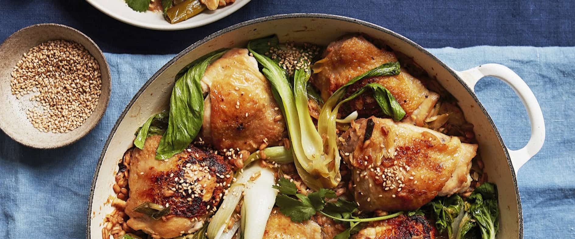 One-Pot-Huhn mit Pak Choi und Perldinkel in einer Pfanne, garniert mit Sesam und frischem Koriander, serviert mit einer Schüssel und Löffel.