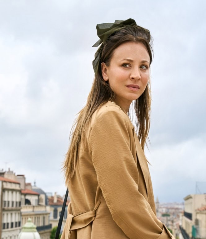 Eine nachdenklich dreinblickende Frau mit Haarschleife steht vor städtischer Kulisse mit historischem Uhrenturm im Hintergrund.