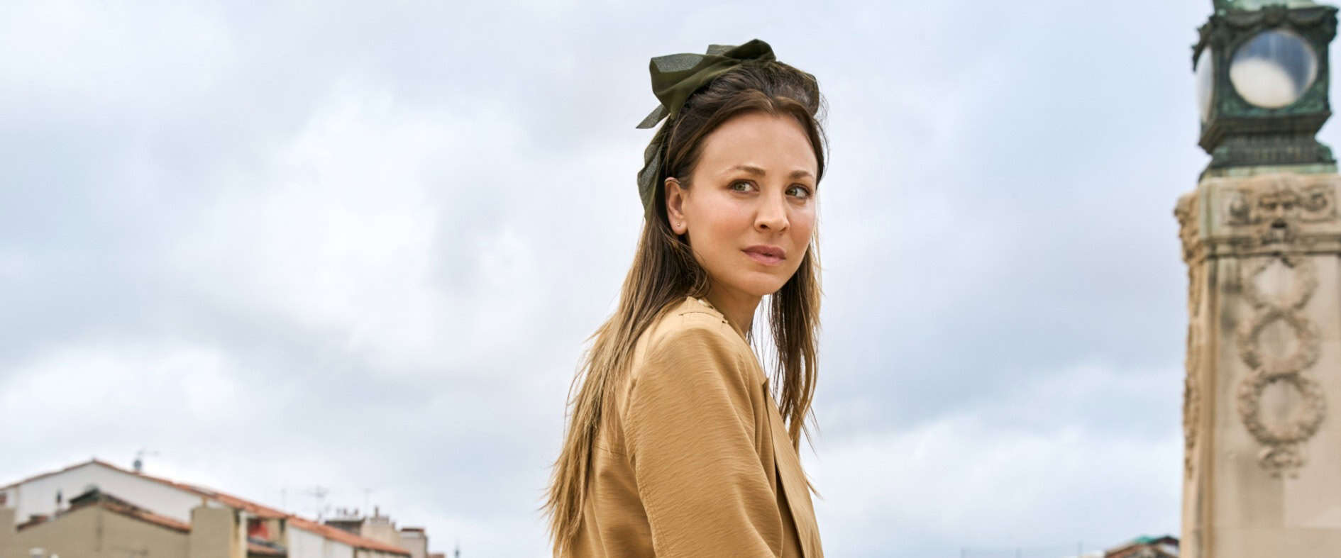 Eine nachdenklich dreinblickende Frau mit Haarschleife steht vor städtischer Kulisse mit historischem Uhrenturm im Hintergrund.