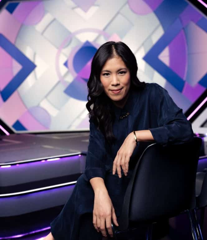 Mai Thi Nguyen-Kim sitzt lächelnd auf einem Stuhl in einem futuristisch gestalteten TV-Studio mit blauen und violetten Lichtelementen im Hintergrund.