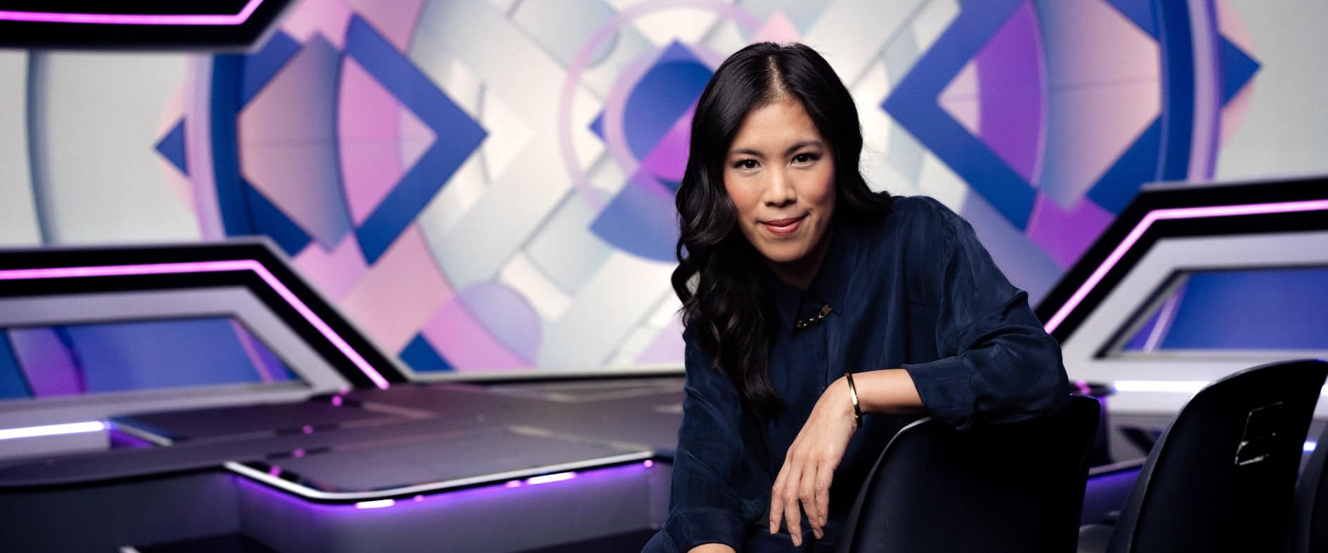 Mai Thi Nguyen-Kim sitzt lächelnd auf einem Stuhl in einem futuristisch gestalteten TV-Studio mit blauen und violetten Lichtelementen im Hintergrund.