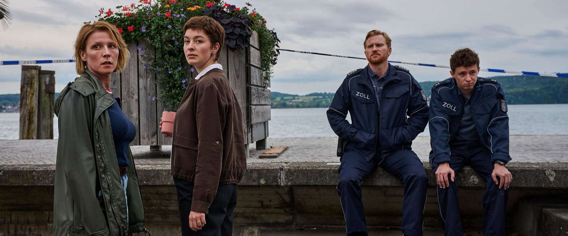 Vier Ermittler aus der ZDF-Krimiserie „Mordufer“ am Bodensee: Zwei Kommissarinnen und zwei Zollbeamte stehen und sitzen konzentriert an einem Tatort mit Blick auf den See und Palmen im Hintergrund.