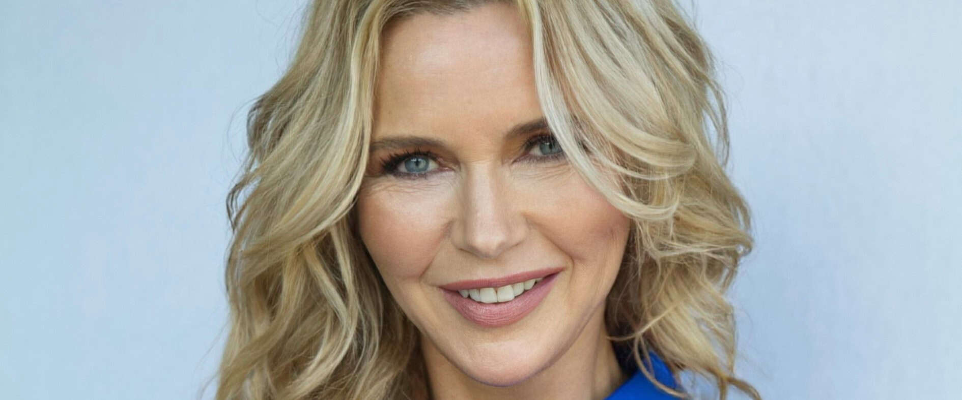 Veronica Ferres, lächelnd und mit verschränkten Armen, trägt eine blaue Bluse und posiert vor hellem Hintergrund.