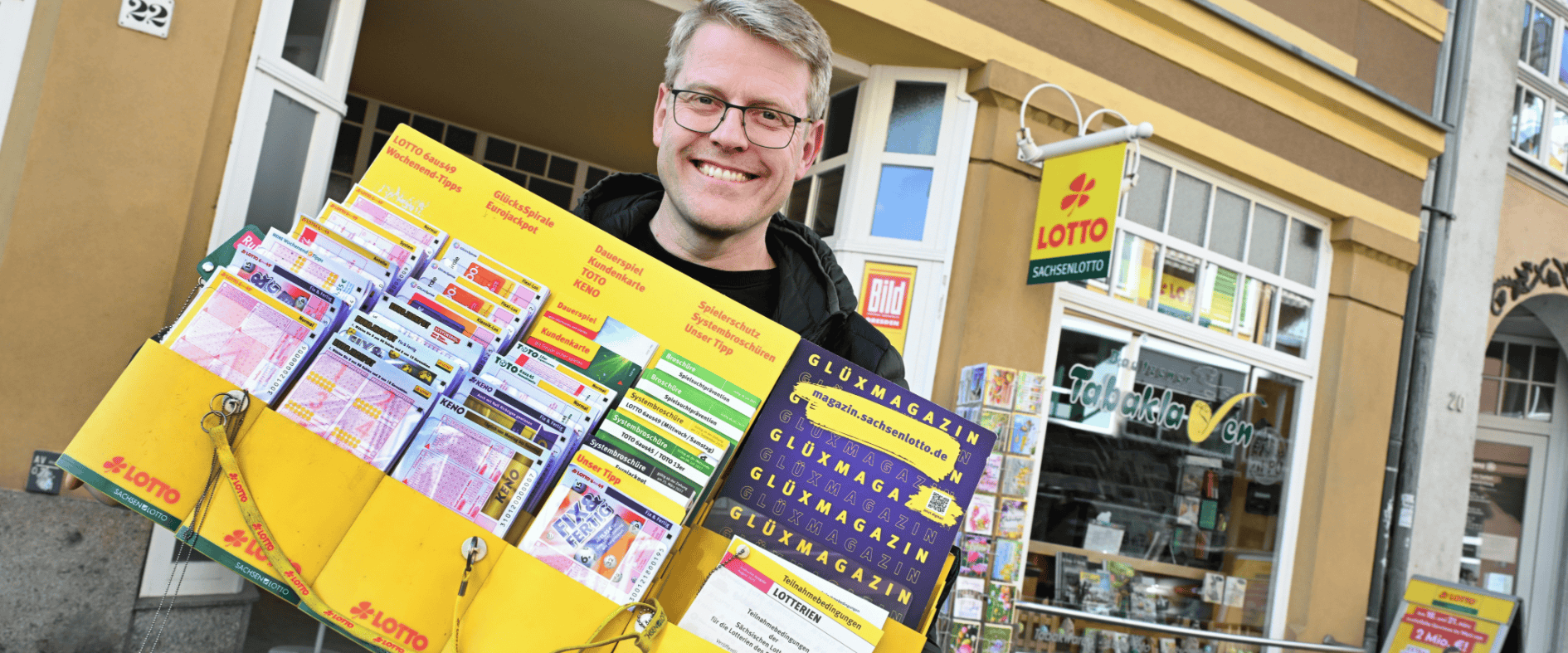 Frank Mietke vor seiner Sachsenlotto Annahmestelle in Bautzen mit einem Präsentationsdisplay für Lotto-Spielscheine, Rubbellose und das glüXmagazin.