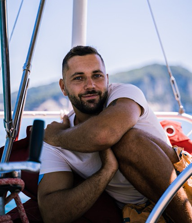 Tim Hund, Abenteurer und Weltumsegler, lächelt entspannt an Bord eines Segelboots mit Blick auf die Küste.