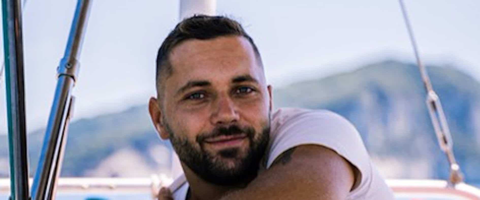 Tim Hund, Abenteurer und Weltumsegler, lächelt entspannt an Bord eines Segelboots mit Blick auf die Küste.