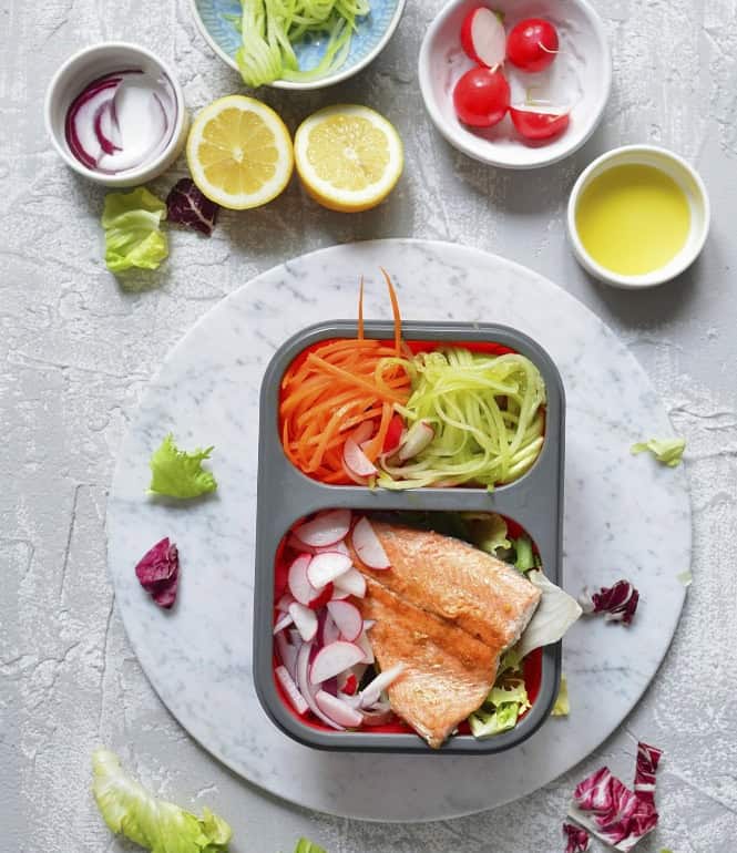 Gesunde Lunchbox mit gebratenem Lachsfilet, Gurken- und Möhrennudeln, Radieschen, Radicchio und Endiviensalat, angerichtet auf einem Marmortisch mit Zutaten wie Zitrone, Olivenöl und Zwiebeln.