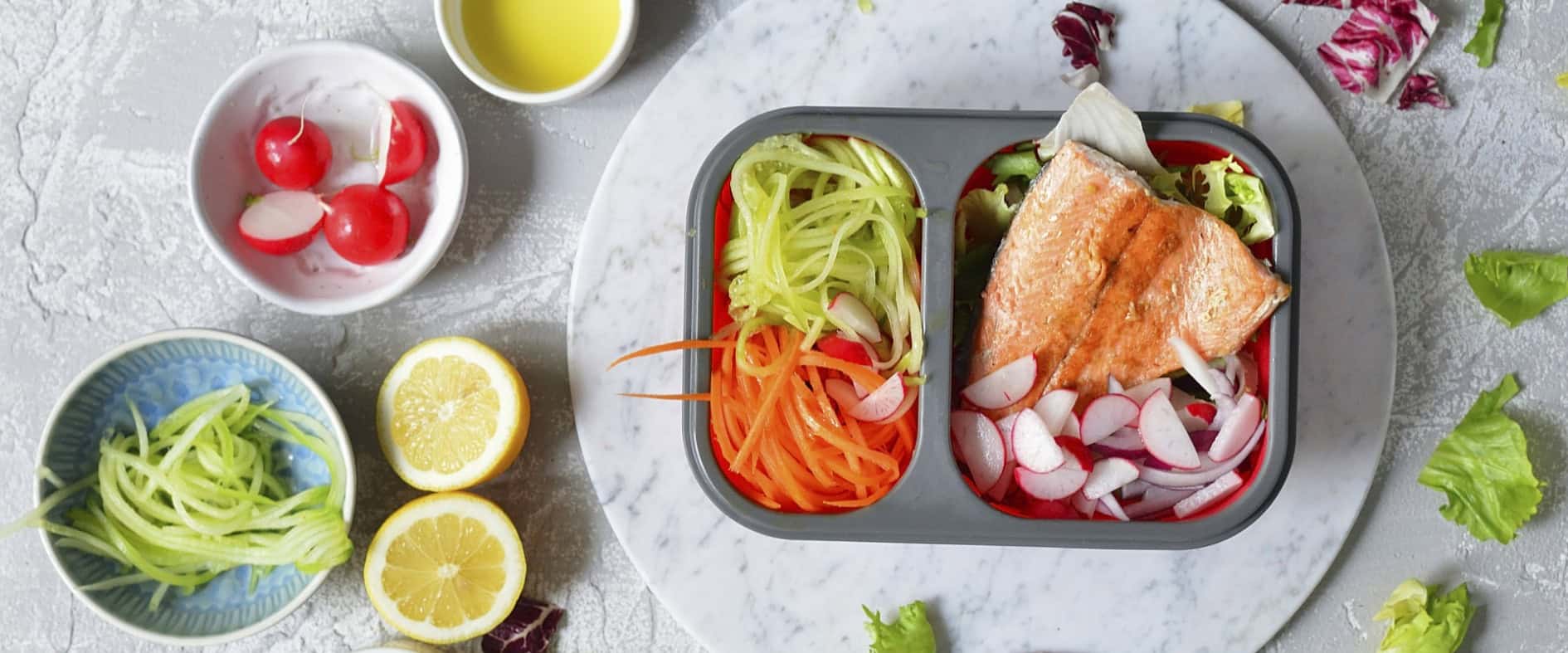 Gesunde Lunchbox mit gebratenem Lachsfilet, Gurken- und Möhrennudeln, Radieschen, Radicchio und Endiviensalat, angerichtet auf einem Marmortisch mit Zutaten wie Zitrone, Olivenöl und Zwiebeln.