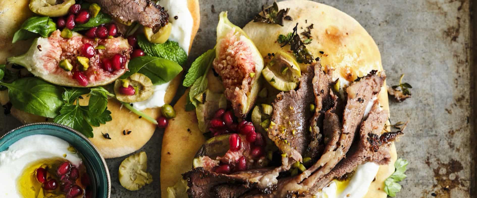 Shawarma-Fladenbrote mit zarten Rindfleischscheiben, Schafsjoghurt, Feigen, Granatapfelkernen, Pistazien und frischen Kräutern auf einem Backblech.