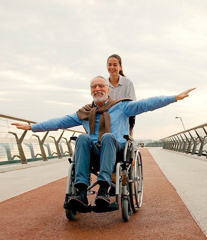 Älterer Mann im Rollstuhl und junge Frau auf einer Brücke, die gemeinsam die Arme ausbreiten und Freude ausdrücken.