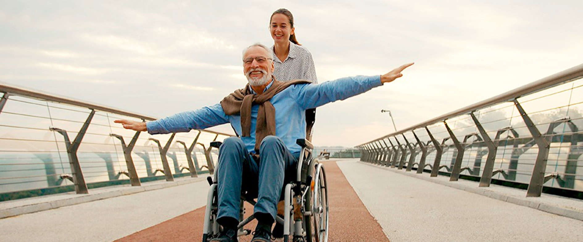 Älterer Mann im Rollstuhl und junge Frau auf einer Brücke, die gemeinsam die Arme ausbreiten und Freude ausdrücken.