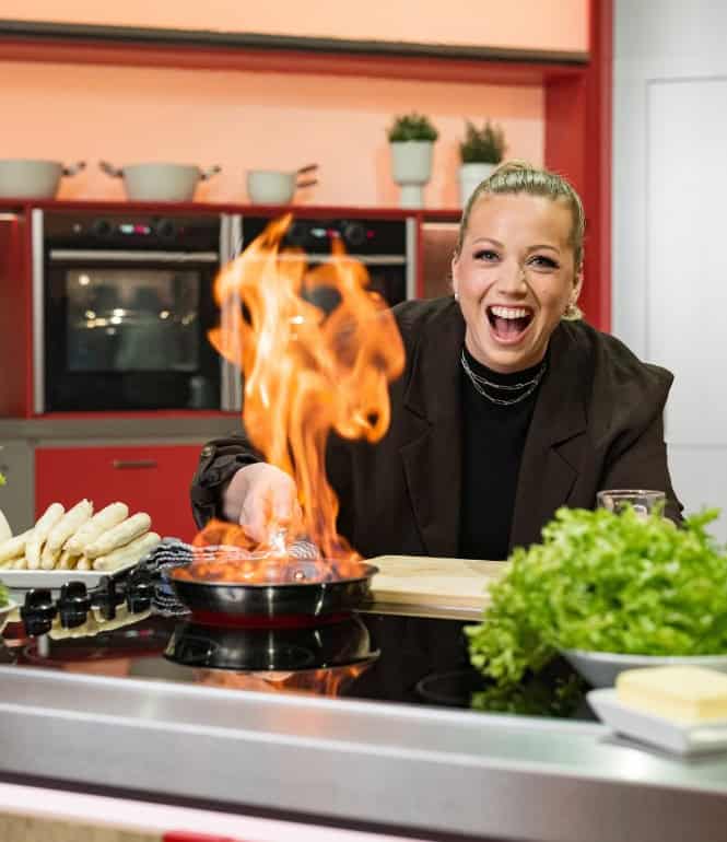 Zora Klipp in Aktion: Die Köchin und Moderatorin der Küchenschlacht XXL flambiert begeistert ein Gericht in einer modernen Küche, umgeben von frischem Gemüse und Kräutern.