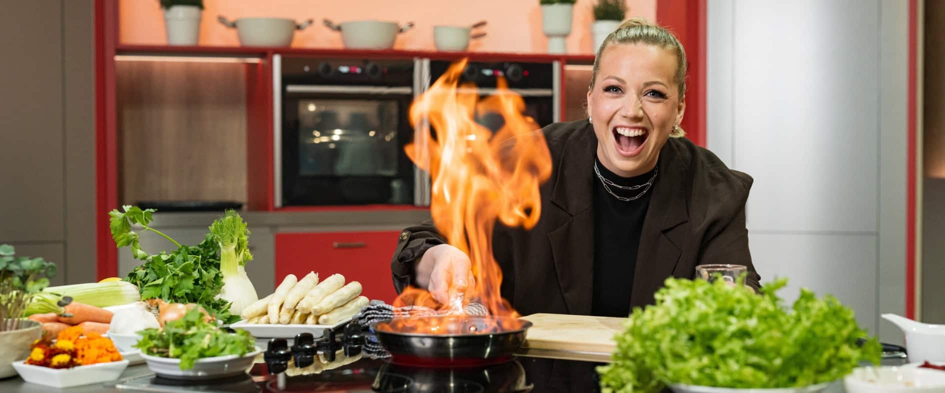 Zora Klipp in Aktion: Die Köchin und Moderatorin der Küchenschlacht XXL flambiert begeistert ein Gericht in einer modernen Küche, umgeben von frischem Gemüse und Kräutern.