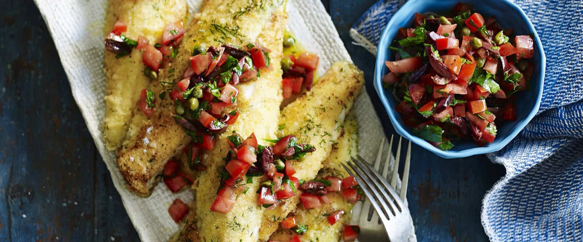 Fischfilets mit Grießkruste und Tomaten-Oliven-Salsa auf einem ovalen Teller, serviert mit Brokkoli und Zucchini sowie einer Schüssel Salsa.